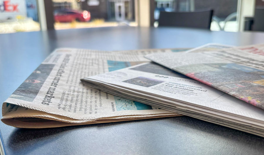Newspapers splayed out on a table, with a street blurred but visible through windows in the background.